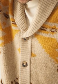 Beige kofta med en ribbad sjalkrage, med ett geometriskt gult mönster och bruna accenter. Inkluderar två runda, mörka knappar.