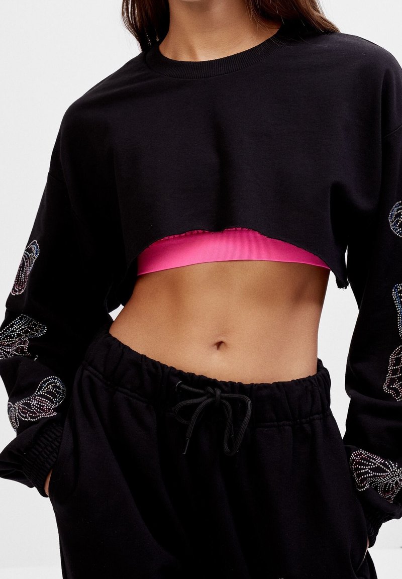 Femme portant un sweat-shirt court noir sur un soutien-gorge de sport rose vif et un pantalon noir à cordon, mains dans les poches, montrant un ventre tonique.