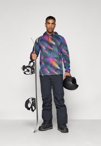 Felpa con cappuccio colorata e a fantasia, con tasca frontale, pantaloni da neve neri, stivali da neve, una tavola da snowboard e un casco nero.