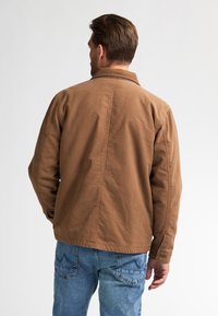 Brown, canvas jacket with a collar, visible seams, and button accents, paired with blue denim jeans.