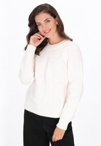 Biały sweter z dzianiny z teksturowanym wzorem w diamenty, okrągłym dekoltem oraz ściągaczami przy mankietach i u dołu. W zestawieniu z czarnymi spodniami.