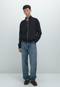 Junger Mann in einer schwarzen Zipjacke, lockeren blauen Jeans und braunen Lederschuhen vor einem schlichten hellen Hintergrund.