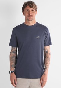 T-shirt en coton bleu marine à manches courtes, col rond, avec un petit logo Oxbow blanc sur la poitrine. Présente des tatouages visibles sur les bras.