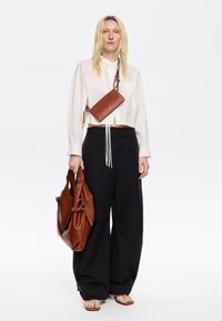 Borse di pelle marrone, una a tracolla con una cintura intrecciata e una borsa oversized. Camicia bianca e pantaloni neri a gamba larga. Sandali bassi.