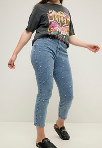 Light blue denim jeans with scattered white pearl embellishments, fitted at the waist, tapering to ankle length, paired with a dark graphic t-shirt.