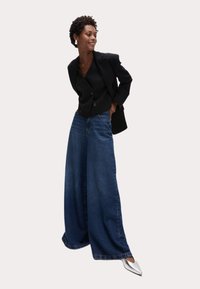 Pantalones vaqueros de pierna ancha en azul oscuro, de cintura alta y con un corte relajado, combinados con un blazer negro ajustado y unos tacones plateados de punta. 