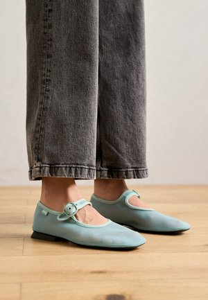 Lyseblå flade sko med spids tå og justerbar rem. Lavet af glat materiale; parret med mørkegrå vidde jeans.