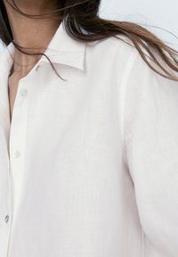 Camicia bianca in lino leggero e morbido, con colletto e bottoni, indossata da una persona con lunghi capelli castani su uno sfondo semplice.