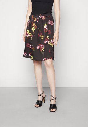 Femme portant une jupe noire fleurie jusqu'aux genoux, un top noir sans manches et des talons ouverts noirs avec des brides à la cheville, debout contre un mur blanc.