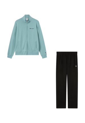 Lichtblauwe Champion-jas met rits en zwarte Champion-sweatpants, beide voorzien van kleine logopatches op mouwen en benen.