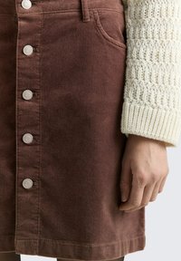 Een bruine denimrok met een voorste knoopsluiting, voorzien van zilveren knopen, zijzakken en een gestructureerde witte gebreide mouw op de achtergrond.