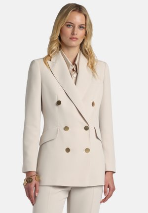 Blazer crem, dublu cu nasturi aurii, revere decupate, buzunare laterale și croială structurată. Poartă peste o bluză deschisă la culoare.