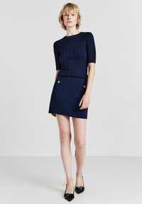 Marineblauer Kabelstrickpullover mit kurzen Ärmeln, kombiniert mit einem marineblauen Minirock mit goldenen Knopfdetails und seitlichen Taschen. Schwarze spitze High Heels.