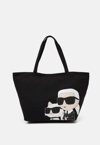 Czarna torba na ramię wykonana z materiału, z rysunkiem w stylu kreskówkowym przedstawiającym postać i kota w okularach przeciwsłonecznych. Szerokie rączki do noszenia.