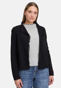 Černý kardigan s třásněmi a texturovaným povrchem, s otevřeným předním dílem a dlouhými rukávy, přeložený přes černo-bílé pruhované tričko.