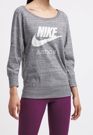 Kobieta ubrana w szarą bluzę Nike z napisem "Just Do It" i fioletowe legginsy, stojąca na tle jednolitej białej ściany.