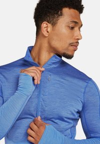 Chaqueta deportiva azul con cremallera, de superficie texturizada, cuello alto y orificios para los pulgares en las mangas; incluye un pequeño bolsillo con cremallera.