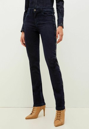 Donkerblauwe denimjeans met een slim-fit ontwerp, voorzien van contrasterende zijnaadstiksels en een medium hoge taille. Gecombineerd met beige hakken met veters.