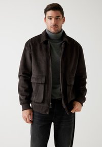 Brun suede bomberjakke med lynlåsfortil, to store frontlommer, ribbede manchetter og en klassisk krave, båret over en mørkegrå turtleneck.