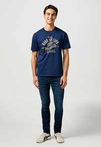 Mörkblå bomull t-shirt med en grafisk tryck som har "Wrangler"-text och mönster. Kombinerad med mörka jeans och ljusa sneakers.