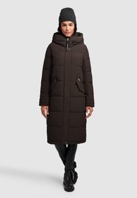 Lang, mørkebrun pufferjakke med hætte og store forlommer. Glat tekstur, quiltet design og lynlåslukning foran. Bæres med en grå beanie.
