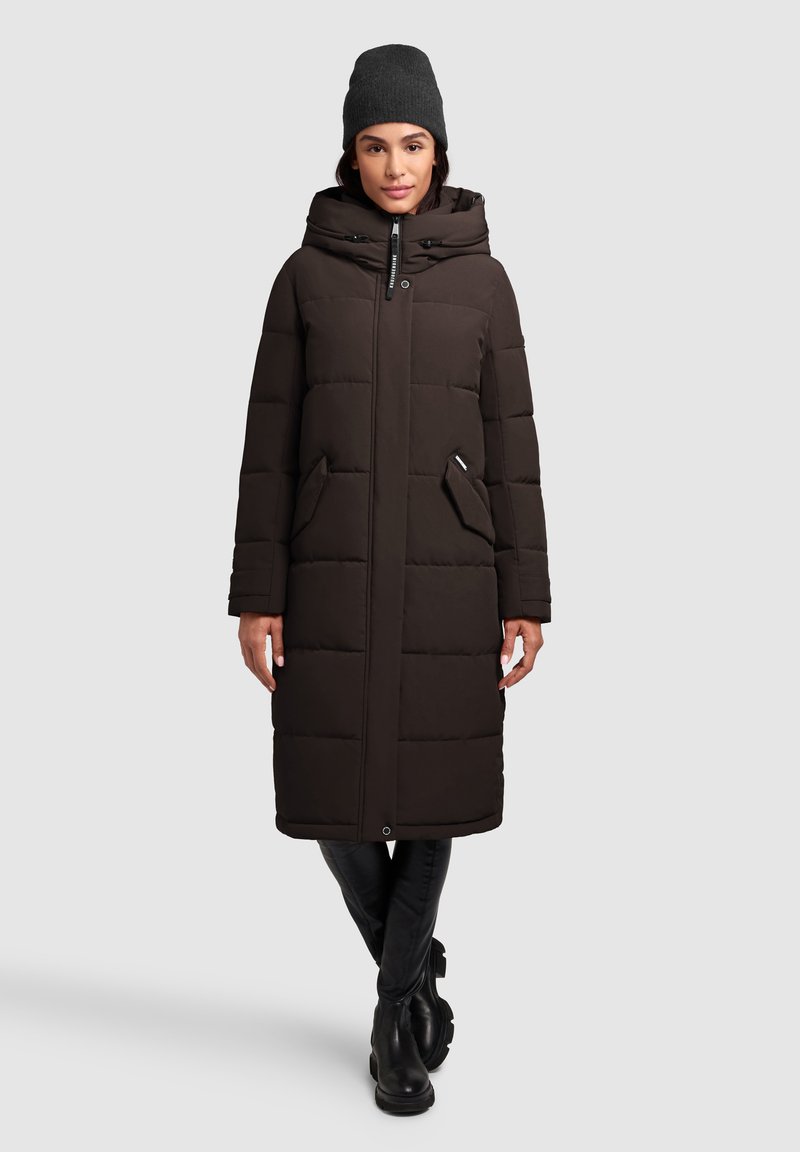 Lang, mørkebrun pufferjakke med hætte og store forlommer. Glat tekstur, quiltet design og lynlåslukning foran. Bæres med en grå beanie.
