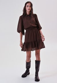 Donna in piedi che indossa un vestito marrone scuro con maniche a sbuffo e orlo a balze, insieme a stivali neri al ginocchio, su uno sfondo chiaro e semplice.