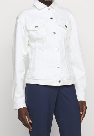 Witte spijkerjas met knopen en twee borstzakken gedragen met donkerblauwe broek bij een persoon met een donkere huid.