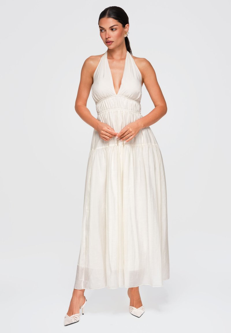 Femme debout portant une robe blanche sans manches à col licou avec taille froncée et des chaussures à talons hauts blanches à bout pointu.
