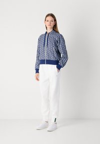 Blå mönstrad zip-up jacka med en figursydd midja och långa ärmar, i kombination med vita sweatpants och minimalistiska vita sneakers.
