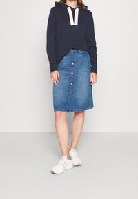 Marinblå hoodie med krämfärgad snörning, kombinerad med en blå denim midi-kjol med framknappar, avslutad med vita sneakers.