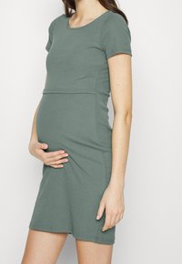 Gravidklänning i ribbat, mjukt grönt tyg med korta ärmar. Den figurnära designen framhäver magen och har en rund halsringning samt knälång fåll.