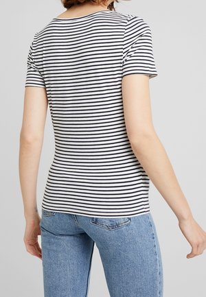 Femme portant un t-shirt rayé horizontal noir et blanc à manches courtes et un jean bleu, vue de dos sur un fond uni.