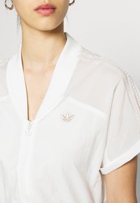 Kobieta ubrana w biały top na zamek z krótkimi podwiniętymi rękawami, prążkowanym kołnierzykiem oraz logo Adidas na piersi, z złotymi kolczykami w kształcie kół.