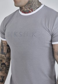 Grå t-shirt med hvid kant, prydet med "SIKSILK" broderet i lys grå på brystet. Lavet af bomuldsmateriale med en blød tekstur.
