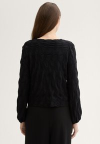 Schwarze blusentexturierte Bluse mit langen Ärmeln, die ein erhabenes Diamant-Muster und einen runden Halsauschnitt aufweist, getragen über dunklen Hosen.