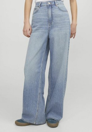 Personne portant un jean large bleu clair et des baskets bleues, debout devant un fond uni.