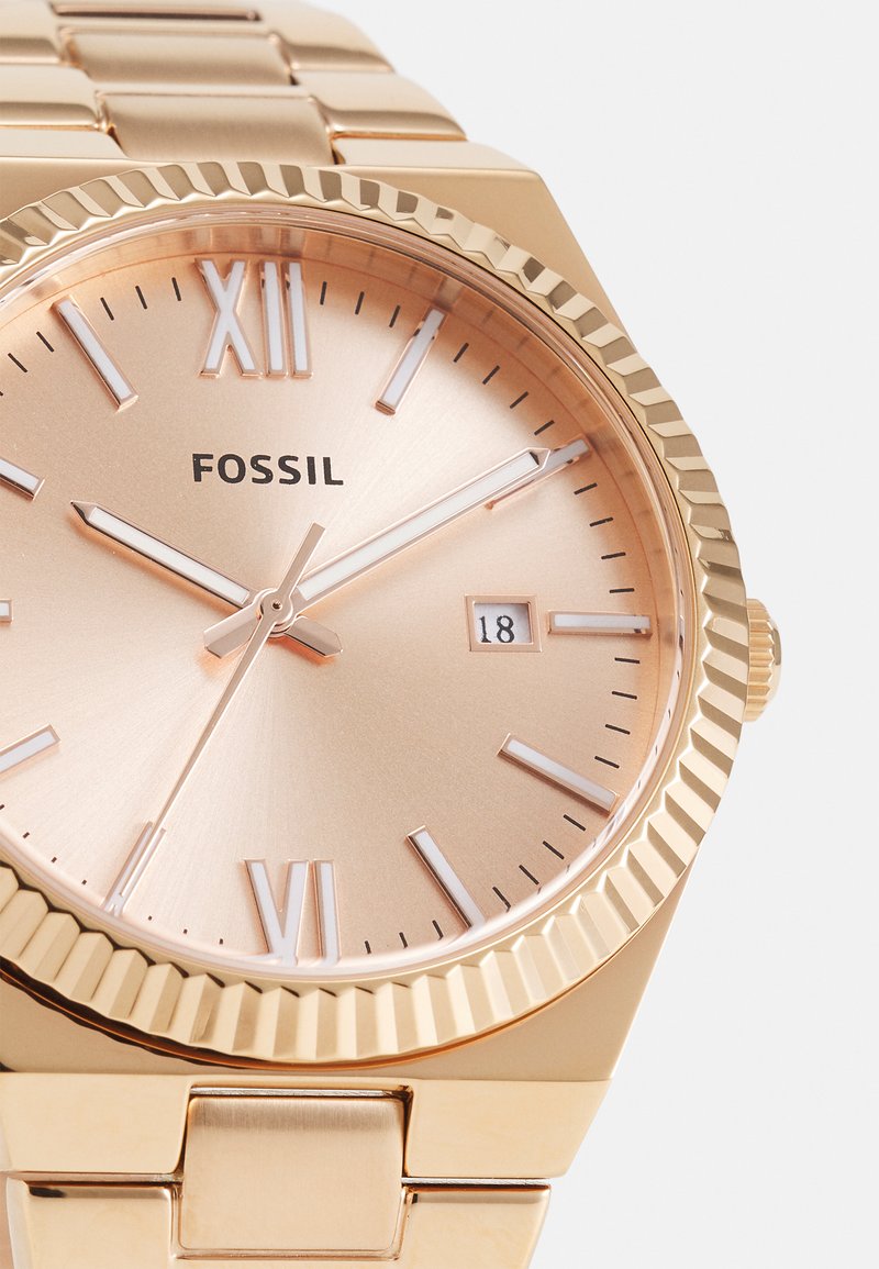 Montre Fossil dorée avec un cadran rose doré, des chiffres romains, une fenêtre de date et une lunette striée, accompagnée d'un bracelet en métal maillé.