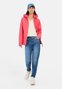 Water-resistant pink jacket with a hood, light beige shirt, and blue jeans. White and gray sneakers complete the outfit.