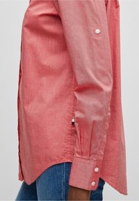 Sidevinkel af en person iført en langærmet rød skjorte med knapper og blå denimjeans.