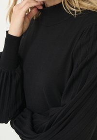 Pull noir à col montant avec manches plissées côtelées et texture lisse. Le tissu est léger et ajusté au poignet.