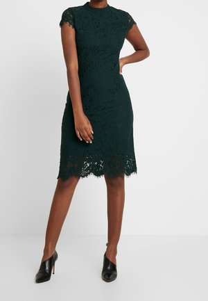 Mujer con un vestido de encaje verde oscuro hasta la rodilla, con mangas cortas y dobladillo festoneado, combinado con zapatos negros de tacón alto.