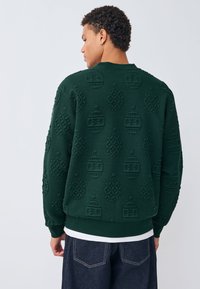 Zielony sweter z fakturą, ozdobiony wypukłymi wzorami, z prążkowanymi mankietami i okrągłym dekoltem, noszony na białej koszuli z ciemnymi dżinsami.