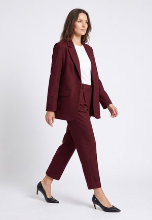 Vrouw loopt, draagt een bordeaux pak met een wit overhemd en zwarte pumps met spitse neuzen op een witte achtergrond.