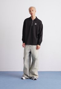 Zwarte longsleeve shirt met een halve rits en wit logo, gecombineerd met lichtgrijze cargobroek met zijzakken en een relaxte pasvorm.