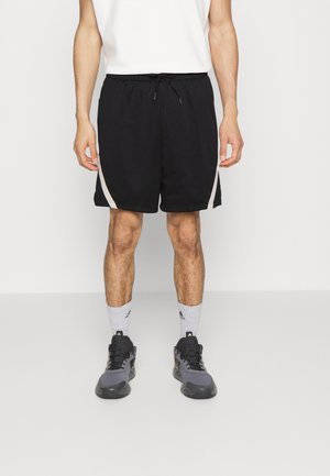 Man die zwarte sportshorts met witte zijkstrepen draagt, grijze en zwarte sportschoenen, witte Adidas-sokken, staand tegen een effen achtergrond.