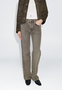 Jeans beige a gamba larga con texture sbiadita, abbinati a una giacca in camoscio marrone scuro e scarpe nere a punta. Design semplice e funzionale.