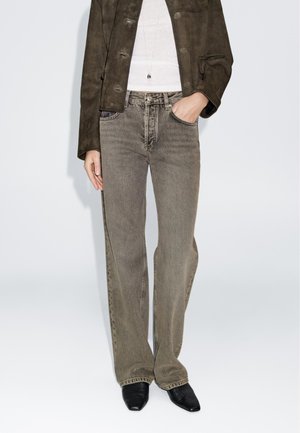 Beige weit geschnittene Jeans mit verwitterter Textur, kombiniert mit einer dunkelbraunen Wildlederjacke und schwarzen spitz zulaufenden Schuhen. Einfaches, funktionales Design.