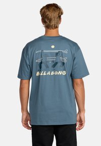 Blå bomull t-skjorte med et grafisk motiv av to personer som bærer surfbrett, med "BILLABONG" tekst under. Korte ermer og avslappet passform.