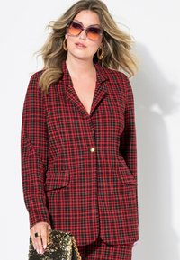 Chaqueta de traje de cuadros rojos y negros con dos bolsillos, solapas en pico, un solo botón dorado y un bolso de lentejuelas doradas, combinada con gafas de sol de gran tamaño.